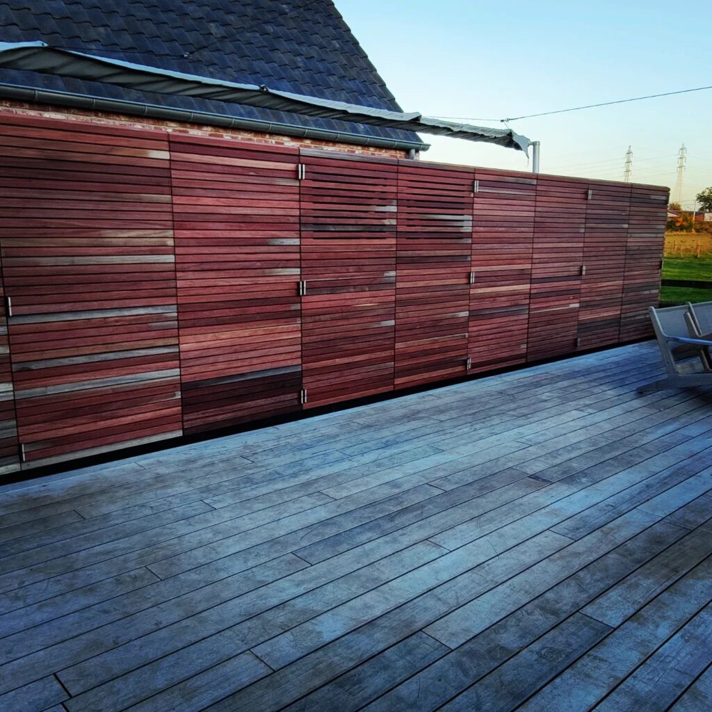 Moderne houten tuinkast met horizontale latten in donkerrood en grijs op een houten terras naast een woning.