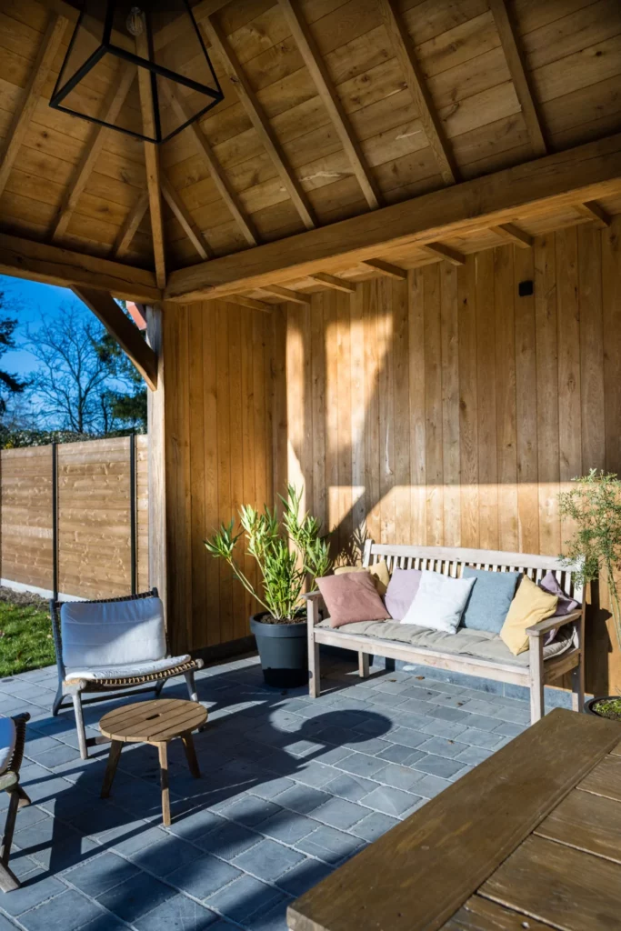 Overdekt terras met houten constructie, witte bank met kleurige kussens, loungestoel, potplanten en houten salontafeltje.